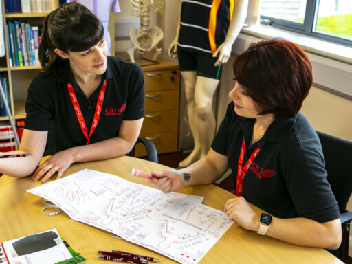 Two members of the DMO clinical team discussing a measurement chart over a wooden table. There is a skeleton and a mannequin in the background. The clinical team are wearing branded black dmo t shirts