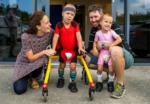 zach and river with their mum and dad outside of the factory in redruth smiling in their DMO suits. River is in his walker