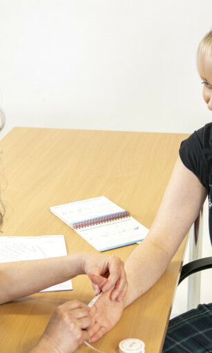 physiotherapist sarah measuring for a glove on a lady with blonde hair. They are both sat at a table