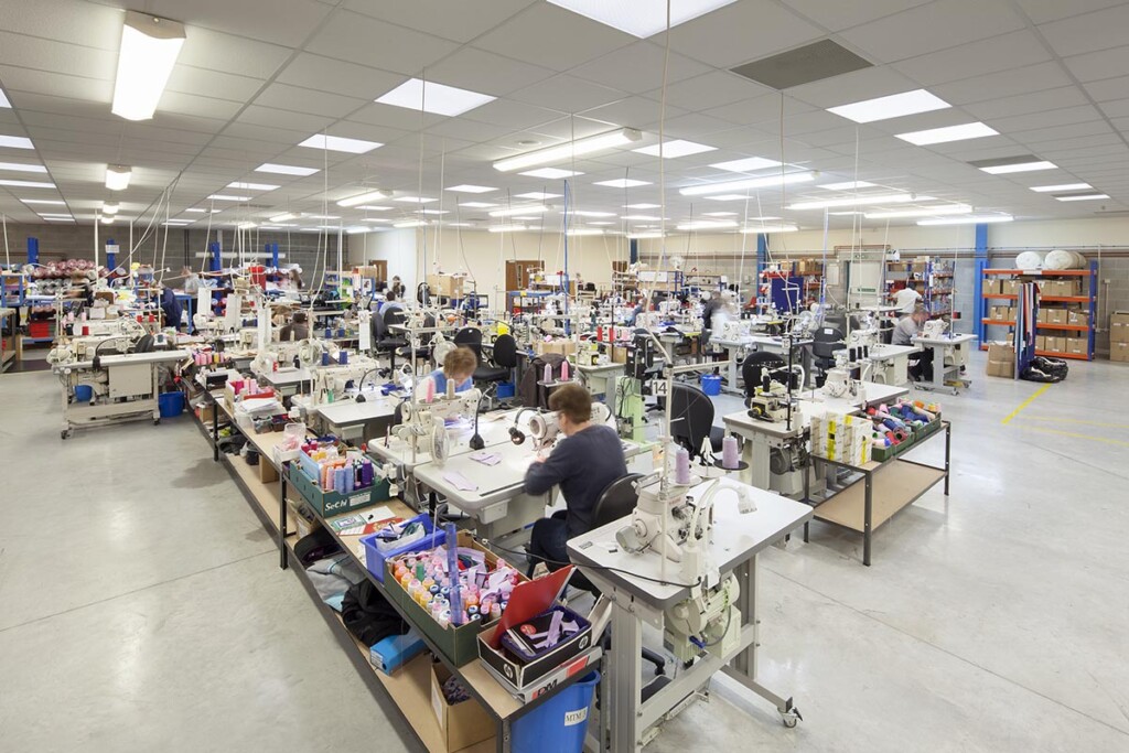 A blurred image of the manufacturing floor. There are rows of machines and cotton reels next to the machines. There are wires hanging down from the ceiling that give the machines power.