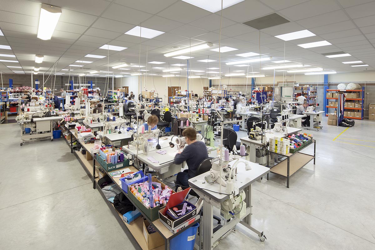 A blurred image of the manufacturing floor. There are rows of machines and cotton reels next to the machines. There are wires hanging down from the ceiling that give the machines power.