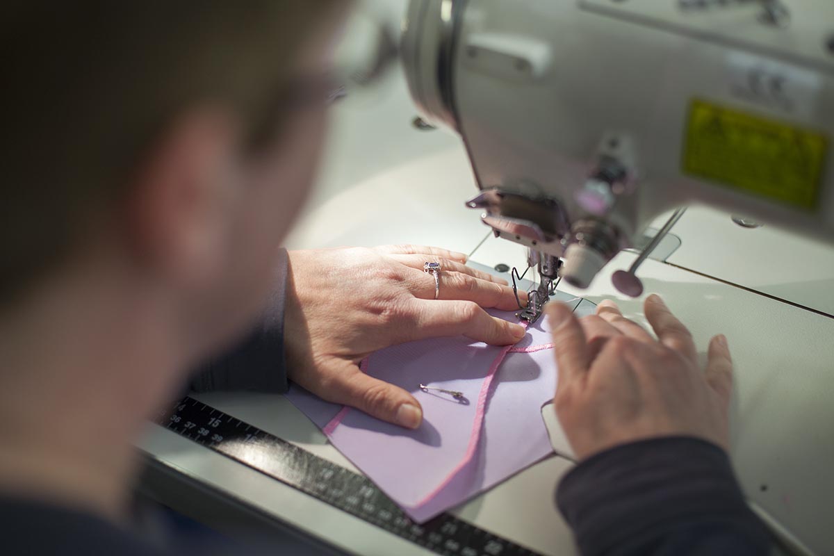 photograph taken over a sewing machinist shoulder where she is machining a lilac orthosis with pink stitching. Her hands are feeding the product through the sewing machine